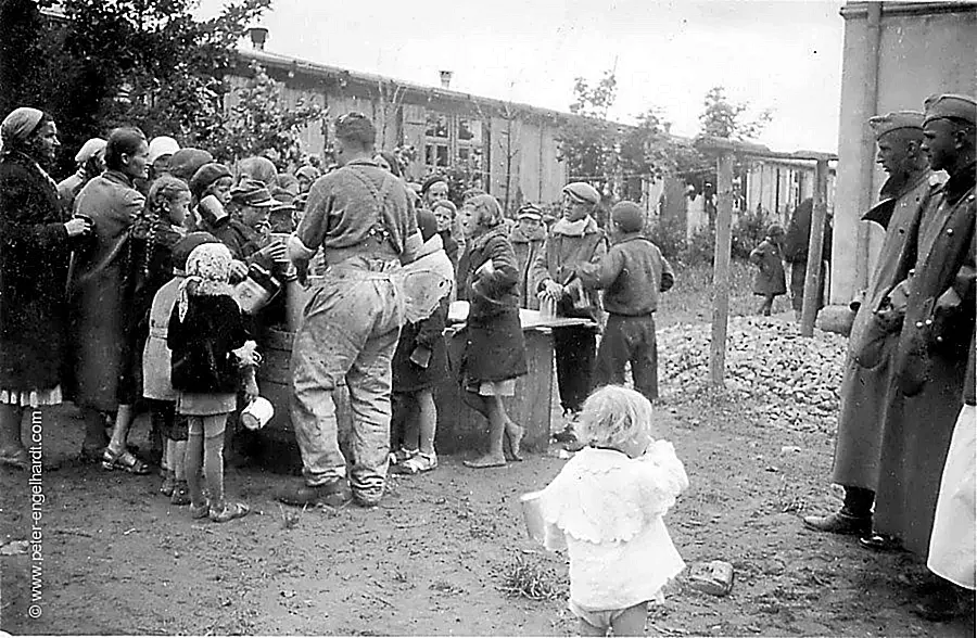 Ein Soldat der Heeresgruppe Nord teilt an eine Gruppe Frauen und Kinder Essen aus, undatiert wohl 1942 bis 1944.